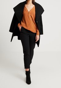 Cappotto nero oversize, blouse arancione con scollatura a V e cinque bottoni, pantaloni neri su misura e stivaletti alla caviglia. Tessuto liscio e silhouette strutturata.