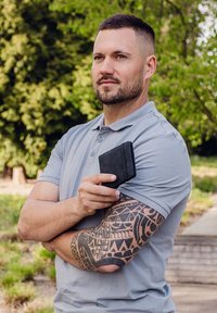 Black wallet held in a hand against a light gray polo shirt. The person has a patterned tattoo on their arm and a blurred green background.
