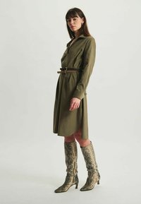 Robe chemise kaki avec un col, taille cintrée par une ceinture brune, associée à des bottes montantes à motif serpent. Tissu de texture lisse.