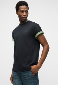 Νεαρός άνδρας που φοράει μαύρο T-shirt με πράσινες ρεβέρ στα μανίκια και μπλε τζιν, στέκεται με το ένα χέρι στην τσέπη και κοιτάζει πλάγια.