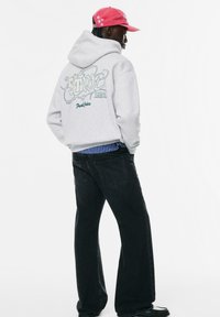 Felpa grigia con grafica sul retro in verde, indossata sopra jeans larghi neri, abbinata a un cappellino rosso con stelle bianche.