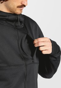 Schwarze Kapuzenjacke aus strukturiertem Stoff, mit hohem Kragen und einer Reißverschluss-Brusttasche. Das Design enthält dezente Rippmuster.