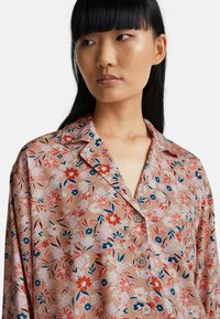 Giovane donna con lunghi capelli neri che indossa una camicia a bottoni floreale beige, che guarda di lato su uno sfondo bianco uniforme.