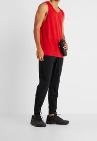 Rød ærmeløs sportstop, sorte sportsbukser og sorte sneakers. Toppen har et diskret logo; bukserne har et strømlinet design.