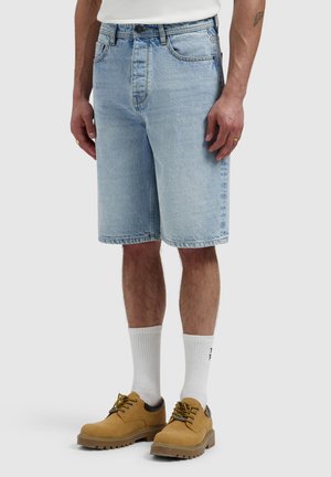Man met lichtblauwe denim shorts, witte sportsokken en lichtbruine veterschoenen, staand tegen een effen lichte achtergrond.