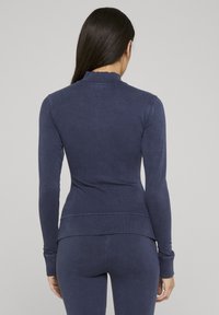 Marineblå langermet topp med en bølget krage, tettsittende design, glatt tekstur og ribbestrikkede mansjetter. Stoffet ser litt slitt ut.