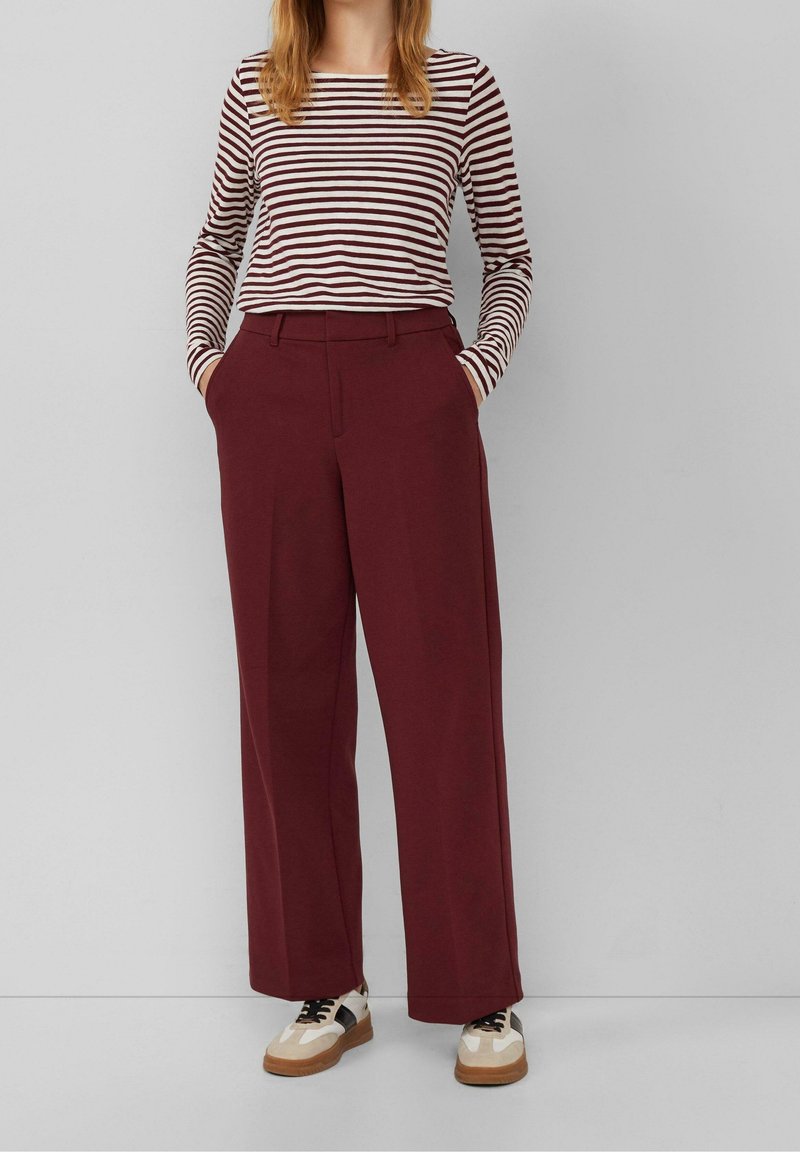 Femme portant un pantalon large bordeaux, un chemisier rayé à manches longues et des baskets blanches avec des accents noirs, les mains dans les poches, debout.