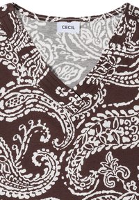 Chemise marron à col en V avec des motifs blancs complexes de paisley et floraux et une étiquette blanche indiquant "CECIL" à l'encolure.