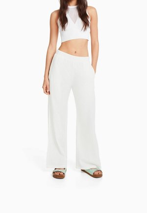 Mujer con pantalones blancos de pierna ancha, top corto blanco con escote transparente y sandalias azul claro con suelas marrones, posando contra un fondo blanco.