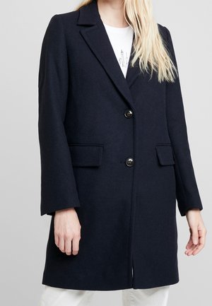 Femme portant un manteau en laine bleu marine foncé longueur genoux avec deux boutons noirs et des poches à rabat sur une chemise blanche.