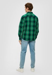 Grünes karierter Hemd mit langen Ärmeln, das kontrastierende schwarze und weiße Linien aufweist, kombiniert mit hellblauen Jeans und weißen Schuhen.