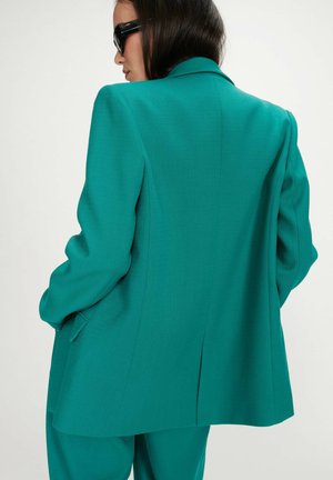 Femme portant un tailleur veste et pantalon turquoise, les mains dans les poches, de profil, portant des lunettes de soleil foncées et les cheveux mi-longs.