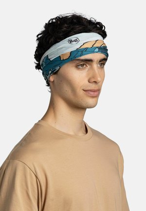 Joven con cabello rizado oscuro, lleva una camisa beige y una cinta para la cabeza Buff con un patrón en azul, marrón y blanco.