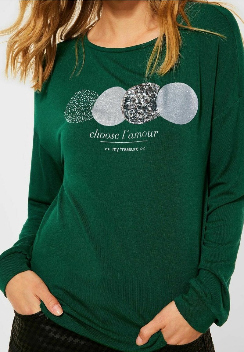 Grön långärmad skjorta med en grafik av glänsande, texturerade cirklar och texten "välja l'amour min skatt" nedanför.