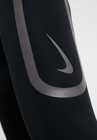 Detailní záběr na černé sportovní legíny s šedým reflexním logem Nike a designem na stehně.