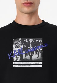 Čierna mikina so grafickým potlačou, ktorá zobrazuje monochromatickú scénu a farebný nápis: "KARL LAGERFELD" v modrej farbe s citátom pod ním.