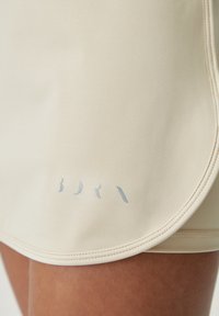 Beige athletic skirt with a smooth texture, featuring a curved hem and subtle silver branding near the bottom edge. Stitched detailing visible.