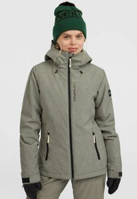 Grijze geblokte skijas met een hoge kraag en ritszakken, gecombineerd met een groene beanie met een zwart logo en een pompon.