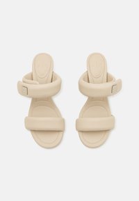 Beige flate sandaler med to brede stropper, en dempet såle og preget logo på siden. Glatt tekstur og minimalistisk design.