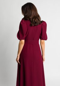 Burgundowa sukienka z bufkowymi rękawami, wcięciem w talii i zwiewnym dołem. Gładka tekstura materiału, elegancki krój z dopasowaną górą.
