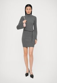 Pilkai dryžuota mezginė suknelė su aukštu kaklu ir ilgomis varpelio formos rankovėmis. Stilingai derinama su grandinine dirželiu ir rožine rankine. Juodi aštriais pirštais aukšti bateliai.