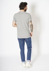 Grå kortærmet t-shirt med klassisk pasform, parret med blå denimbukser og hvide sneakers. Synlige tatoveringer pryder armen.