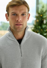 Pull en tricot gris avec un col montant et une fermeture éclair. Présente un tissu texturé avec un design à côtes. L'arrière-plan inclut un sapin de Noël flou.