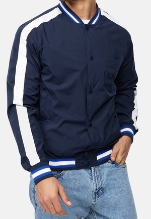 Mężczyzna noszący granatową kurtkę bomber z białymi paskami na rękawach i mankietami w niebiesko-białe paski, w połączeniu z jasnoniebieskimi dżinsami.