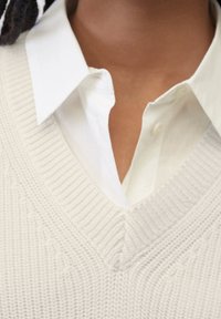 Ljus beige stickad tröja med V-hals över en vit skjorta med krage. Tröjan har ett strukturerat mönster och ribbade kanter.