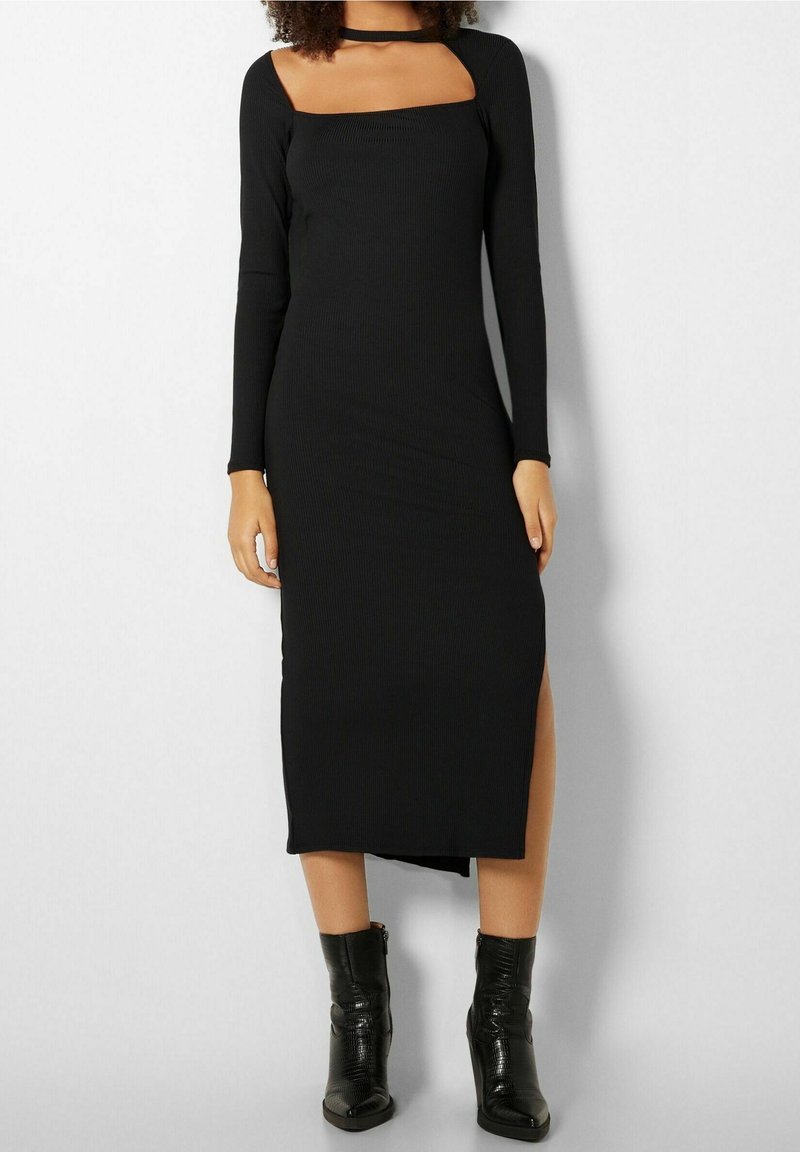 Femme portant une robe midi noire à manches longues avec une encolure carrée découpée et des bottines noires à talon léger.