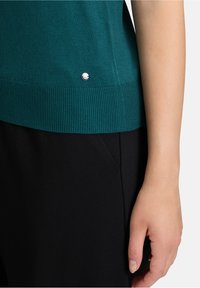 Turf groene gebreide trui met geribde onderrand, voorzien van een decoratieve zilveren accent, gecombineerd met zwarte stoffen broek. Close-up van hand en mouw.
