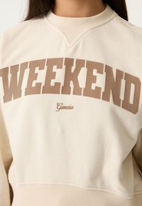 Sudadera beige con la palabra "WEEKEND" en grandes letras marrones, con un cuello acanalado y textura suave. Logotipo "Garcia" en una fuente más pequeña debajo.
