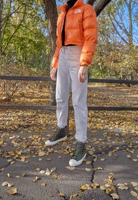 Pomarańczowa kurtka puchowa, czarny golf, jasnoszare spodnie i zielone modne trampki za kostkę z białymi podeszwami, w jesiennym otoczeniu na zewnątrz.
