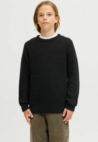 Schwarzer Strickpullover mit strukturierendem Muster, Rundhalsausschnitt und langen Ärmeln. Über einem weißen Hemd getragen, kombiniert mit olivgrünen Hosen.