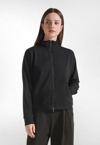 Donna che indossa una giacca nera con zip e colletto alto, con pantaloni scuri a righe sottili, che guarda leggermente di lato con una mano in tasca.