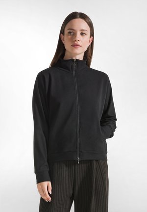 Vrouw in een zwarte jack met rits en hoge kraag, met een donkere krijtstreepbroek, die iets opzij kijkt met één hand in haar zak.