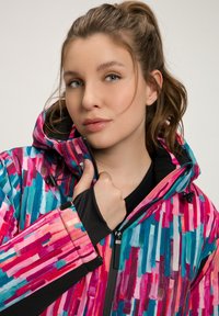 Giacca colorata e decorata con strisce verticali in rosa, blu e verde acqua. Include una chiusura con cerniera sul davanti e un design con cappuccio. Tessuto testurizzato visibile.