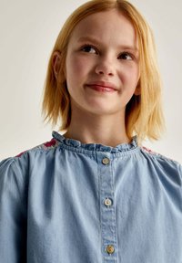 Cămașă din denim bleu deschis, cu guler plisat și broderie florală pe umeri. Are închidere cu nasturi și croială lejeră.