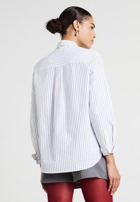 Camicia bianca con bottoni, caratterizzata da sottili righe verticali grigie, dotata di yoke posteriore, maniche arrotolabili e orlo curvo.