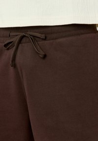 Pantalon de survêtement marron avec une taille à cordon de serrage, texture côtelée et finition lisse, avec une vue rapprochée du matériau.