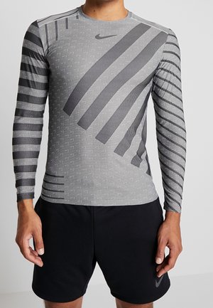 Hombre con camiseta deportiva de manga larga gris con patrones diagonales y rayas negras, combinada con pantalones cortos negros con logotipos de Nike.