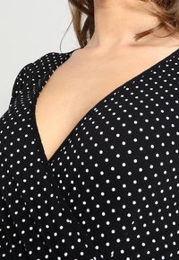 Svart topp i tyg med prickar, med v-ringning. Har vita prickar och draperad design, vilket ger ett mjukt, strukturerat utseende.