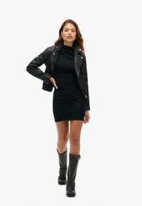 Veste de biker en cuir noir sur une robe noire moulante à fronces, associée à des bottes noires à genoux avec des boucles.