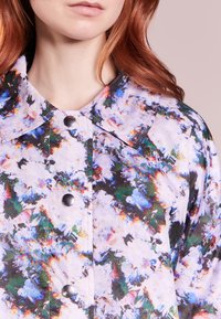 Langærmet skjorte med et blomsterprint i lilla, grønne og blå nuancer. Den har en stor krave og sølvfarvede trykknapper ned foran.