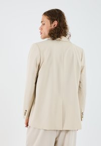 Blazer taillé beige au design structuré, à manches longues et avec des poignets à trois boutons ; coutures minimales ; tissu lisse et léger.