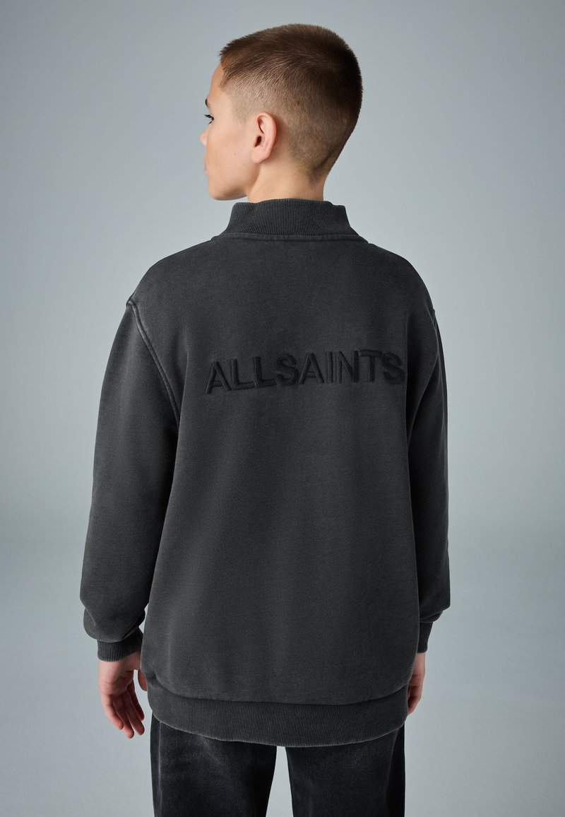 Junge Person mit kurzen Haaren, die einen dunklen Sweatshirt mit "ALLSAINTS" auf dem Rücken trägt, seitlich vor einem einfarbigen Hintergrund.