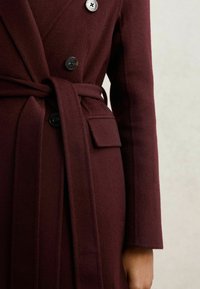 Burgundy uldfrakke med dobbeltbrystet design, der har sorte knapper, et bælte og synlige sidelommer. Blødt, struktureret stof.