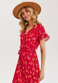 Czerwona sukienka w kwiaty z dekoltem w serek, krótkimi falbaniastymi rękawami i wiązanym paskiem w talii. Posiada wzory w żółte i różowe kwiaty na lekkim materiale.