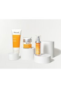 Murad hudvårdsprodukter visas på vita cirkulära baser. Funktioner inkluderar en tube, en burk och en pumpflaska, alla med orangea accenter.