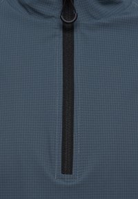 Nærbillede af en mørkegrå jakke front med en lodret sort lynlås og en rund trække-flig på tekstureret stof med gittermønster.